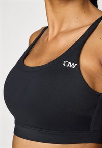 Soutien-gorge de sport noir avec un décolleté arrondi, de larges bretelles et une coupe ajustée. Inclut un petit logo blanc sur le côté gauche. Tissu lisse.