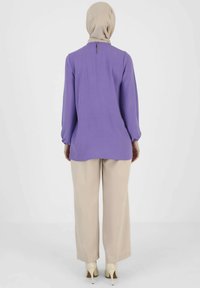 Purple, long-sleeve tunic with a high neckline and back keyhole detail, paired with beige wide-leg trousers and cream heels.