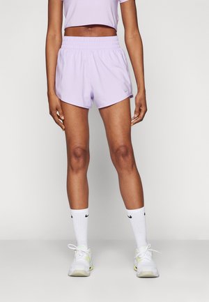Femme portant un short de sport violet clair et un crop top assorti, des chaussettes blanches Nike et des baskets blanches, debout devant un fond uni.
