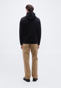 Homme debout, de dos, portant un sweat à capuche noir, un pantalon beige et des chaussures noires sur un fond blanc uni.