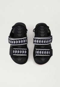 Černé sandály s otevřenou špičkou a dvěma nastavitelními pásky, s opakujícím se vzorem bílého loga, vystavené vedle sebe na jednobarevném pozadí.