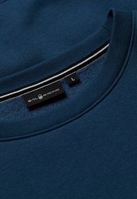 Sudadera azul marino con cuello redondo acanalado, que presenta una etiqueta negra con "SAIL RACING" y tamaño "L," acentuada por rayas blancas.