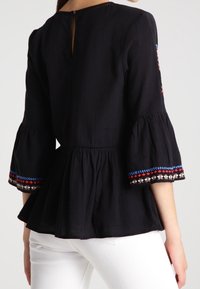 Top peplum nero con maniche a campana, caratterizzato da vivaci dettagli ricamati ai bordi e una apertura a chiave sul retro. Tessuto testurizzato.