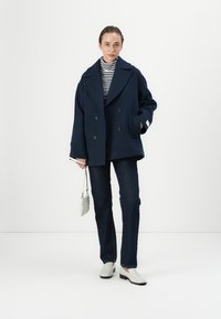 Cappotto pea in lana blu navy con bottoni doppi, abbinato a un dolcevita a righe, jeans scuri, scarpe bianche e una borsa leggera.