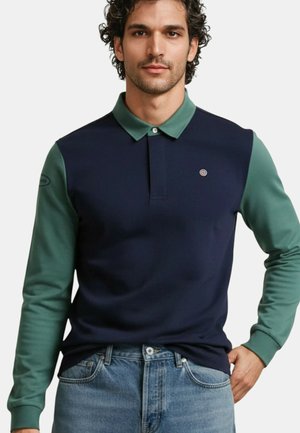 Homme portant un polo à manches longues bleu marine et vert avec col et manches verts, rentré dans un jean bleu, main dans la poche.