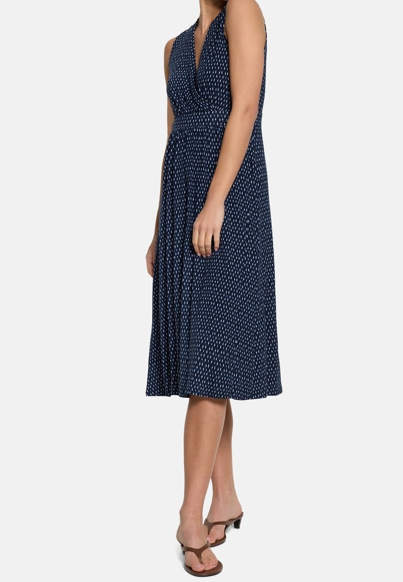 Femme portant une robe midi bleu marine sans manches avec un motif à pois blancs et des sandales à talons marron, debout devant un fond uni.