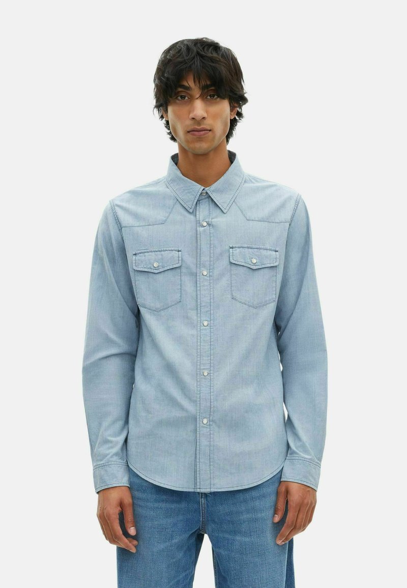 Camicia in denim azzurro chiaro con colletto BUTTON-DOWN, due tasche sul petto e cuciture in evidenza; presenta maniche lunghe e un orlo curvo.