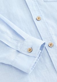 Lyseblå hørskjorte med knapforside, med brune knapper på manchet og stolpe, og blødt, tekstureret stof.