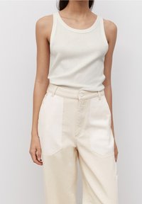 Femme portant un débardeur crème sans manches et un pantalon beige bicolore taille haute avec de grandes poches avant sur un fond uni.