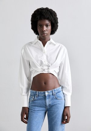 White cropped shirt with button closure and metal accents, paired with light blue denim jeans. Smooth texture, tailored fit, and structured sleeves.