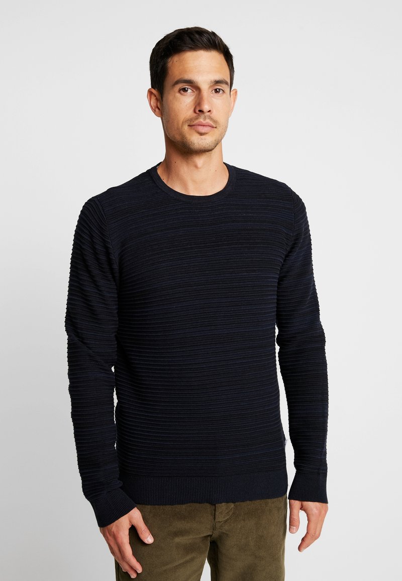 Maglione a coste blu navy con maniche lunghe e scollatura rotonda, caratterizzato da un motivo testurizzato e polsini aderenti, abbinato a pantaloni verde oliva.