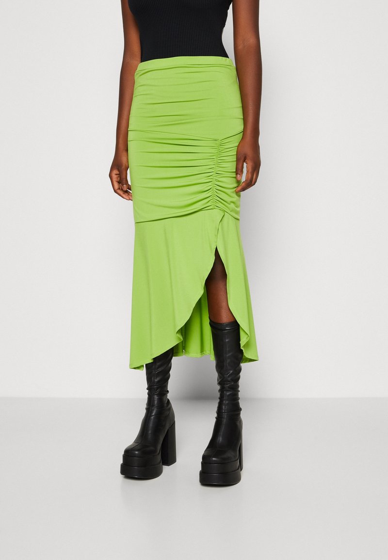 Jupe midi verte froncée avec un ourlet asymétrique, en tissu extensible, dotée d'une fente latérale et associée à des bottes noires à talons épais.