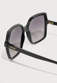 Černé sluneční brýle s odvážným geometrickým designem, které mají tmavé gradientní čočky a zlaté logo "Calvin Klein" na boční straně rámu.