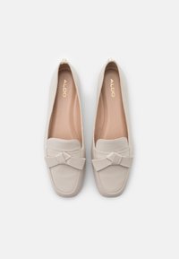 Mocassins en cuir beige avec un nœud décoratif sur l'avant, forme arrondie, texture lisse et détails de logo dorés à l'intérieur.