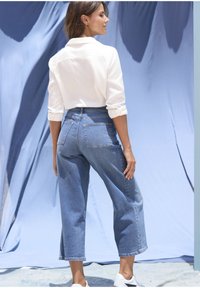 Weißes Hemd mit aufgerollten Ärmeln, kombiniert mit hoch taillierten, weit geschnittenen blauen Jeans aus Denim mit Gesäßtaschen und ausgefransten Säumen.