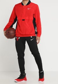 Giacca pullover Nike rossa con accento nero, tasca frontale con patta e colletto blu, abbinata a pantaloni jogger neri con logo dei Bulls rosso.