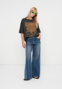 Zwarte oversized t-shirt met oranje grafisch ontwerp, gecombineerd met wijde blauwe denim jeans en groene zonnebril. Ontspannen pasvorm en casual stijl.