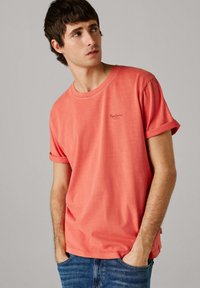 Mladý muž v koralovom tričku s vyhrnutými rukávmi a modrých džínsoch sa pozerá do strany s rukami v vreckách na jednoduchom pozadí.