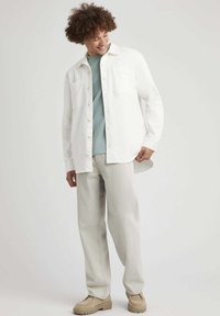 Chemise blanche à boutons, portée sur un haut texturé bleu clair, associée à un pantalon gris clair et des chaussures beiges ; elle dispose de deux poches poitrine.
