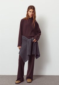 Maglione a costine marrone scuro con collo alto, abbinato a pantaloni a gamba larga. Un coprispalle grigio lavorato a maglia è annodato in vita. Scarpe beige slip-on.