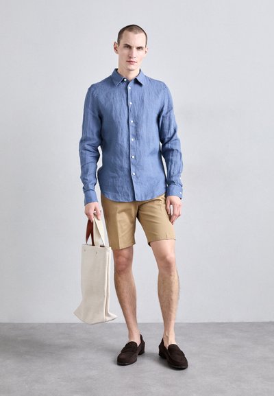 Camisa de lino azul claro con botones, pantalones cortos beige, zapatos de ante marrón sin cordones, y una bolsa de lona crema con asas marrones.