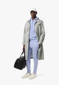 Grau karierter Trenchcoat, helllila Kapuzenpullover und passende Hose, weiße Mütze, hält eine schwarze Tasche, beige Sneaker mit Akzenten.