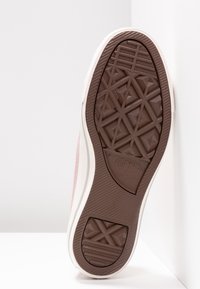 Semelle en caoutchouc marron avec motif géométrique de la bande de roulement sur une basket blanche et rose inclinée contre des surfaces blanches.