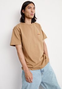 Brown cotton t-shirt with a relaxed fit, featuring a small embroidered logo in red and yellow on the chest, paired with light blue jeans.