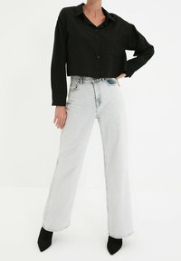 Osoba nosząca czarną, krótką koszulę na guziki, jasne, wysokie, szerokie dżinsy i czarne, szpiczaste botki na obcasie, stojąca z ręką na biodrze.