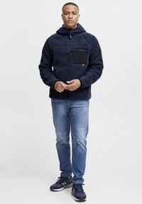 Pullover in pile di colore navy con tasca a marsupio, chiusura a mezza zip, superficie testurizzata e tasca sul petto nera. Indossato con jeans blu e scarpe da ginnastica nere.