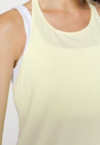 Donna con una canottiera bianca e un top senza maniche di un tenue giallo pallido, che mostra la spalla e la parte superiore del torace.