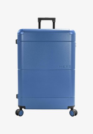 Blauwe koffer met harde schaal, telescopisch handvat en vier dubbele zwenkwielen, rechtopstaand op een witte achtergrond.