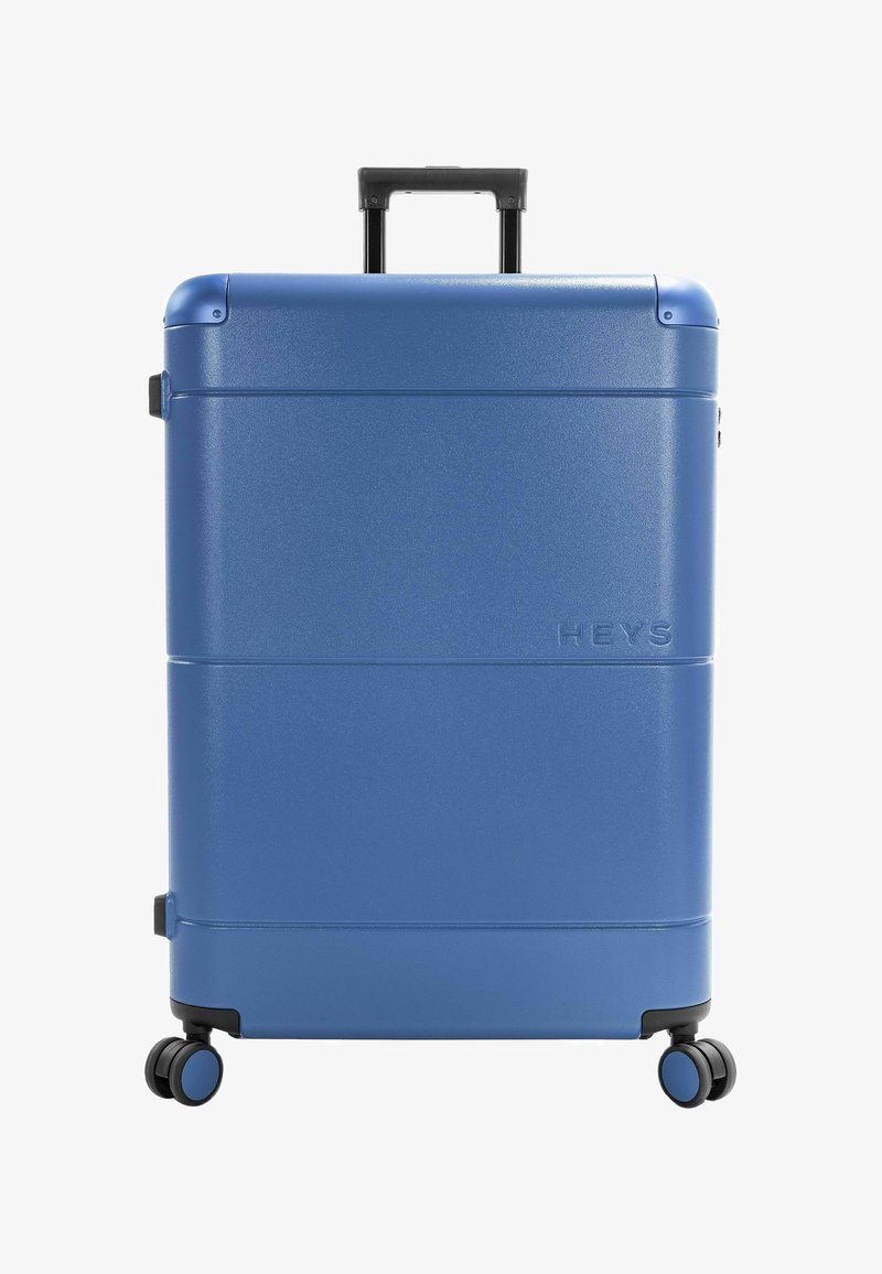 Valise rigide bleue avec poignée télescopique et quatre roulettes doubles pivotantes, posée verticalement sur un fond blanc.