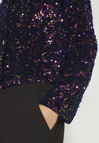 Haut à manches longues bleu foncé recouvert de sequins multicolores associé à un pantalon noir, une main posée négligemment dans la poche.
