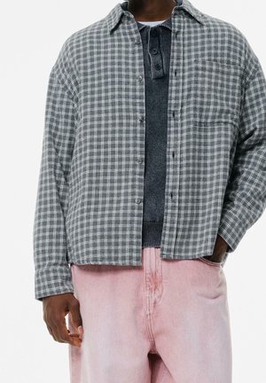 Personne portant une chemise à carreaux grise à boutons par-dessus un pull gris foncé et un pantalon rose clair, une main dans la poche, sur fond blanc.