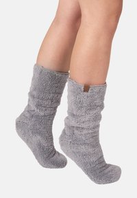 Calcetines grises, peludos y hasta la rodilla con una superficie texturizada y un pequeño detalle de cuero en la parte superior. Diseñados para ofrecer calidez y comodidad.