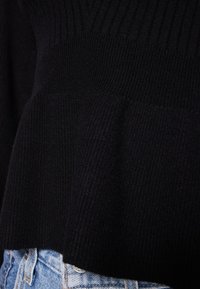 Pull noir en maille avec texture côtelée, présentant une coupe ample et un design superposé ; présenté avec un jean en denim clair.