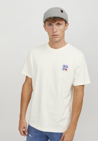 Weißes Baumwoll-T-Shirt mit rundem Halsausschnitt, kurzen Ärmeln und einem blau-roten rechteckigen Logo auf der linken Brust. Lässige Passform, simples Design.