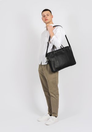 Sac en cuir noir avec double anses supérieures, surface texturée et une bandoulière ajustable, tenu par une personne en chemise blanche et pantalon beige.