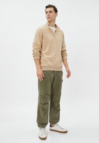Sudadera cremosa con cremallera y puños acanalados, combinada con pantalones cargo holgados en color oliva. Zapatillas blancas con suelas marrones completan el conjunto.