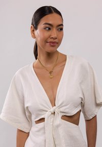 Weißes Leinenoberteil mit kurzen Ärmeln, tiefem V-Ausschnitt und geknoteter Front. Goldene Kettenhalskette mit einem runden Anhänger am Hals.