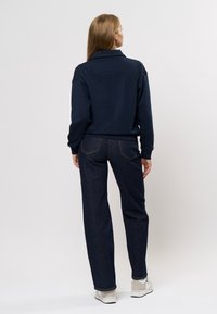 Marinegenser med høy krage, avslappet passform og lange ermer, sammen med mørk denimbukse med vide ben og hvite sneakers.