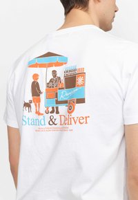 Witte T-shirt met een grafisch ontwerp op de achterkant met een voedselwagen, karakters in bruin, blauw en oranje, en de tekst "Stand & Deliver."