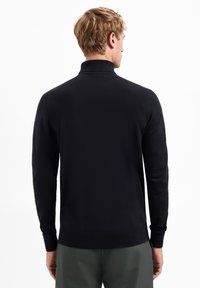 Zwarte longsleeve polo trui met hoge kraag, gebreide textuur en aansluitende manchetten, gecombineerd met groene broek, gezien vanuit de achterkant.