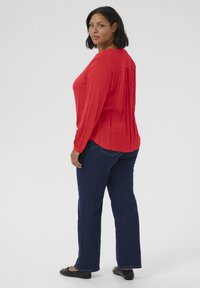 Blusa rossa a maniche lunghe con scollatura rotonda, abbinata a jeans dritti blu scuro e ballerine nere, su uno sfondo neutro.