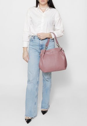 Vrouw die een witte blouse en lichtblauwe spijkerbroek draagt, met een grote roze leren handtas vastgehouden aan de handvatten.