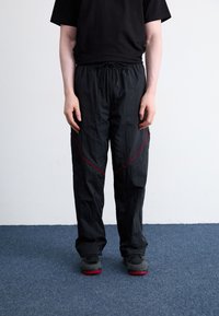 Zwarte lichtgewicht broek met een tailleband met koord, voorzien van contrasterende rode diagonale stiksels en een relaxte pasvorm. Gecombineerd met zwarte schoenen.