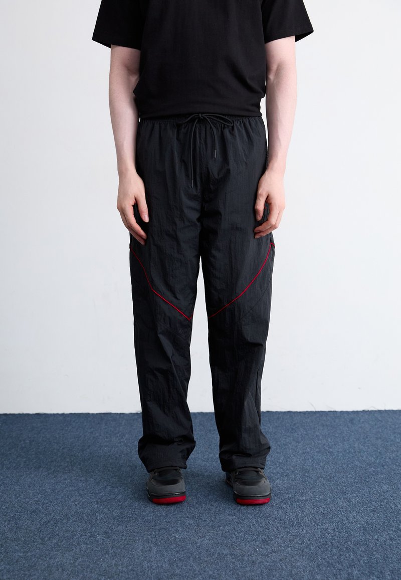Zwarte lichtgewicht broek met een tailleband met koord, voorzien van contrasterende rode diagonale stiksels en een relaxte pasvorm. Gecombineerd met zwarte schoenen.