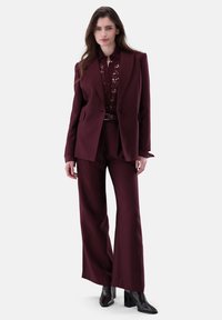 Completo su misura bordeaux con blazer doppiopetto e pantaloni a gamba larga, abbinato a una blusa in pizzo e stivali neri con tacco.