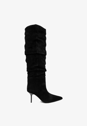 Bota alta hasta la rodilla de ante negro con diseño holgado, punta puntiaguda y tacón de aguja delgado de altura media, vista lateral sobre fondo blanco.